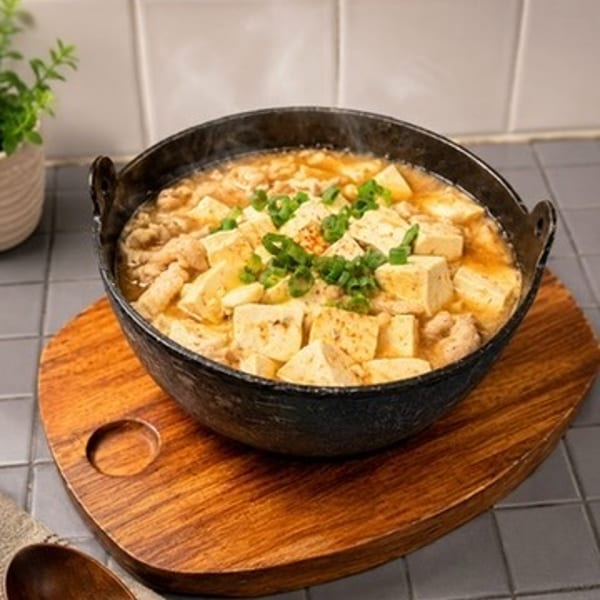 Steaming hot tofu and meat stew in a black pot on a wooden trivet.