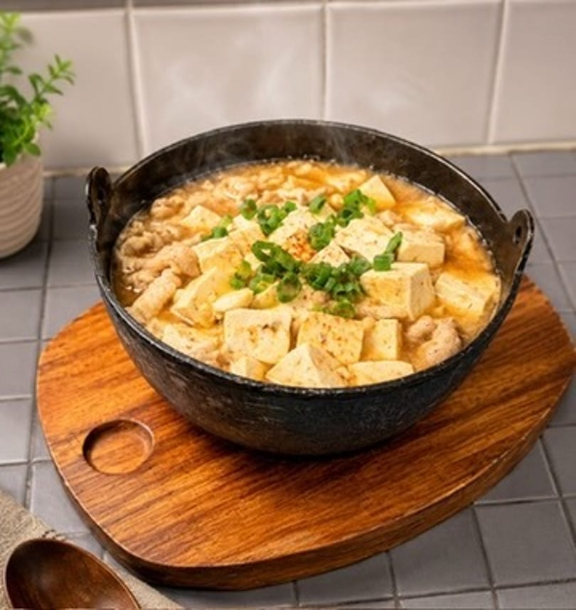 Steaming hot tofu and meat stew in a black pot on a wooden trivet.